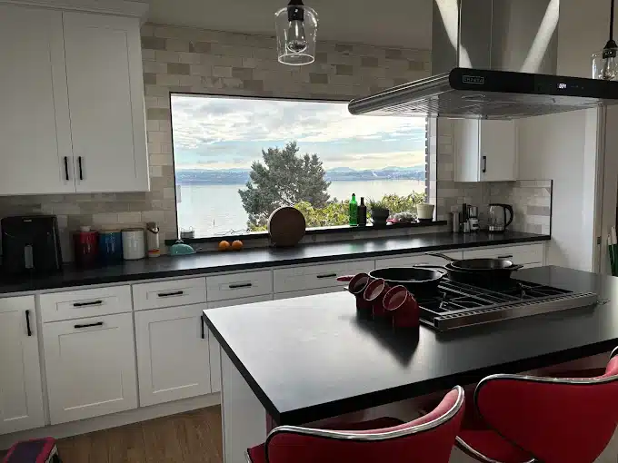 Modern white kitchen with black countertops, large window, and scenic waterfront view.